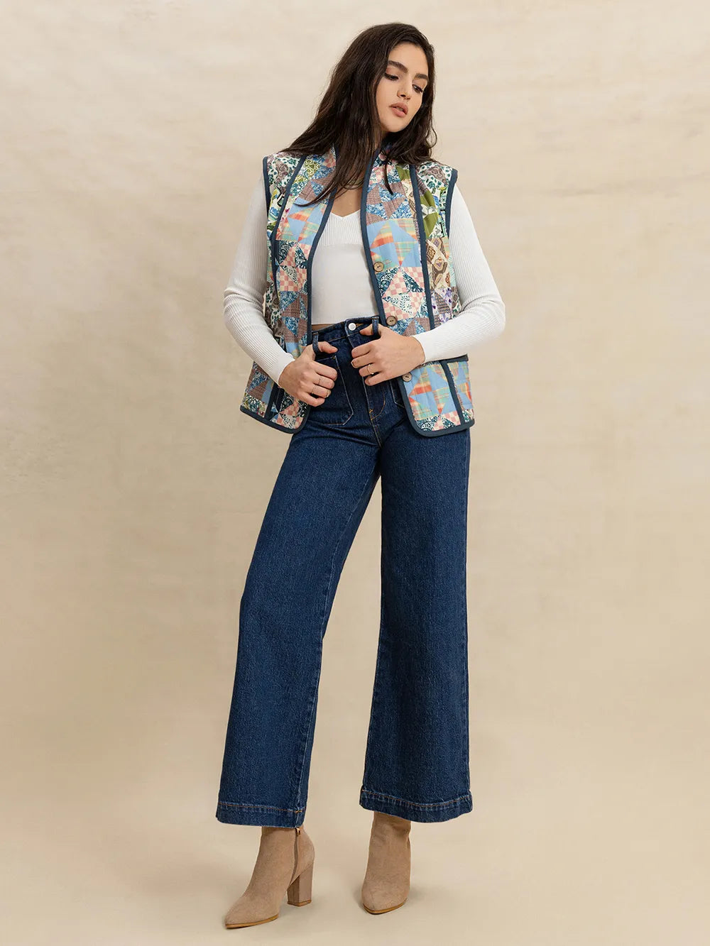 Woman wearing a floral patterned vest over a white top and blue jeans on a beige background