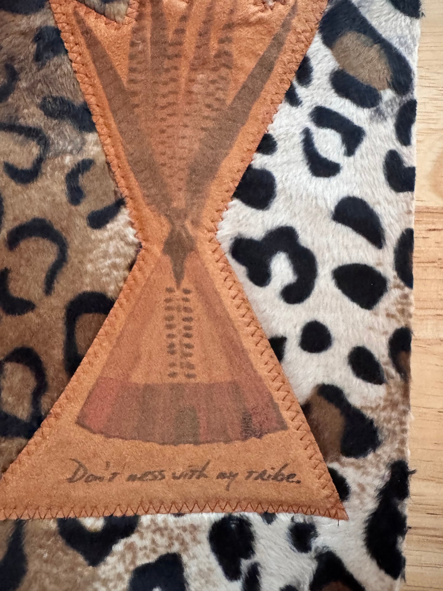 A close-up of a leopard-print fabric item, likely a blanket or rug, featuring a central tan stitched geometric patch with a hand-drawn teepee and the handwritten text 'Don't mess with my tribe.'.