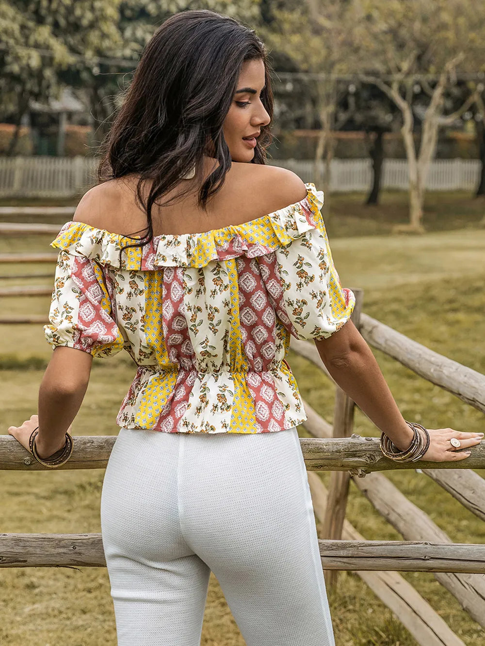 Woman wearing a colorful off-shoulder top and white pants standing near a wooden fence in a park.