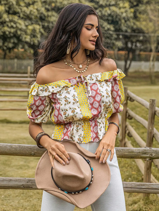 Woman wearing a colorful off-shoulder top holding a brown hat in an outdoor setting.
