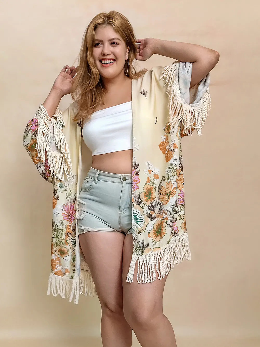 Woman wearing a floral kimono with fringe, white top, and denim shorts on a beige background