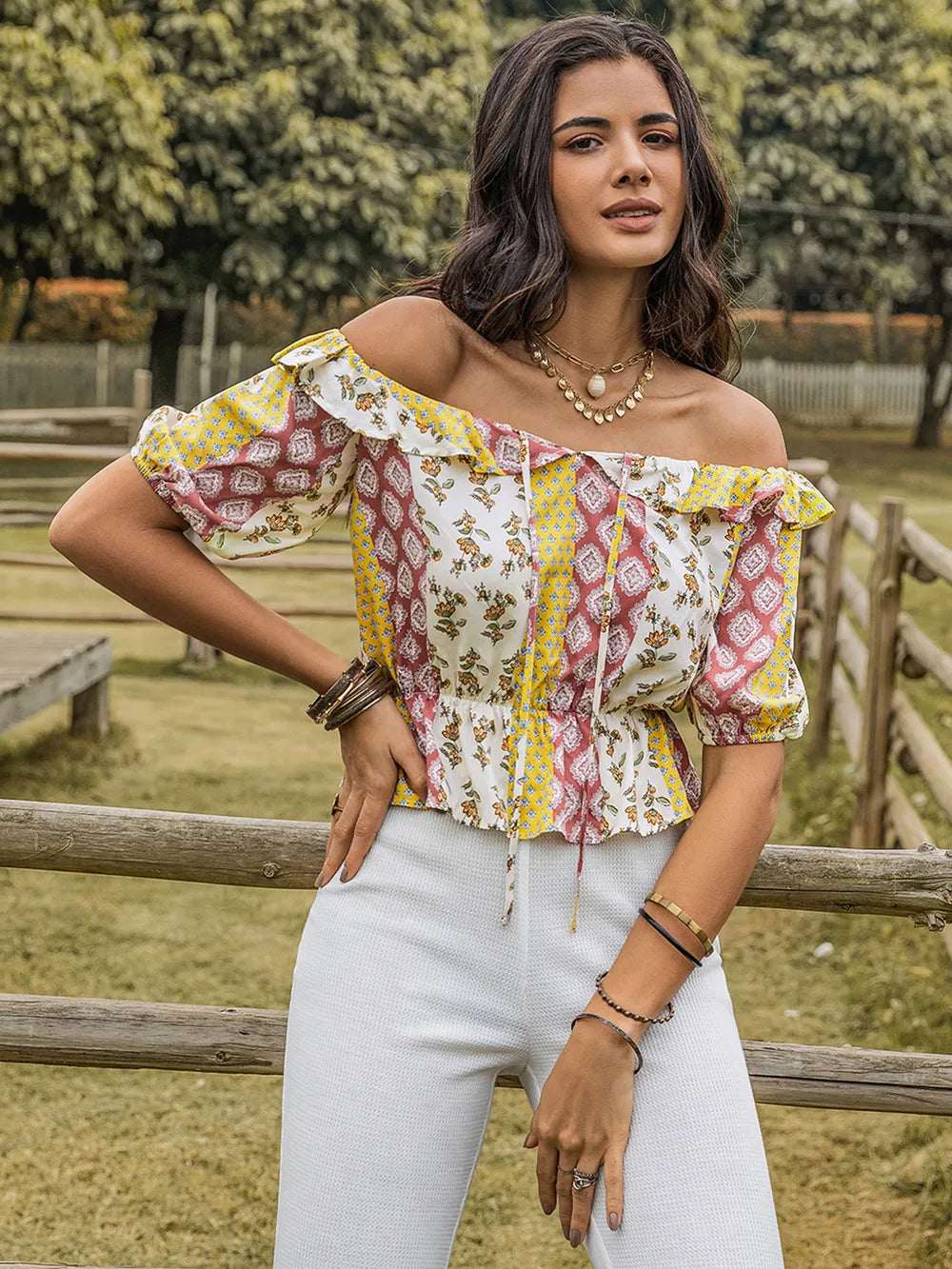 Woman wearing a colorful off-shoulder top and white pants standing outdoors with a wooden fence and trees in the background.