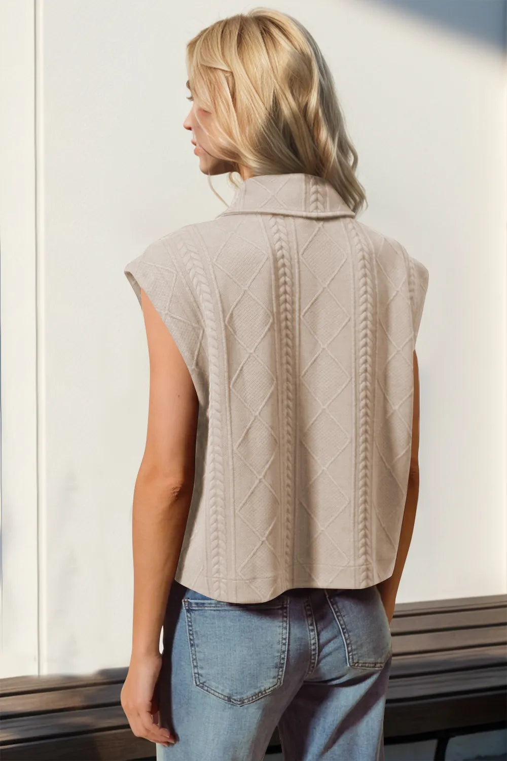 Woman wearing a beige knitted vest and blue jeans standing against a light background.
