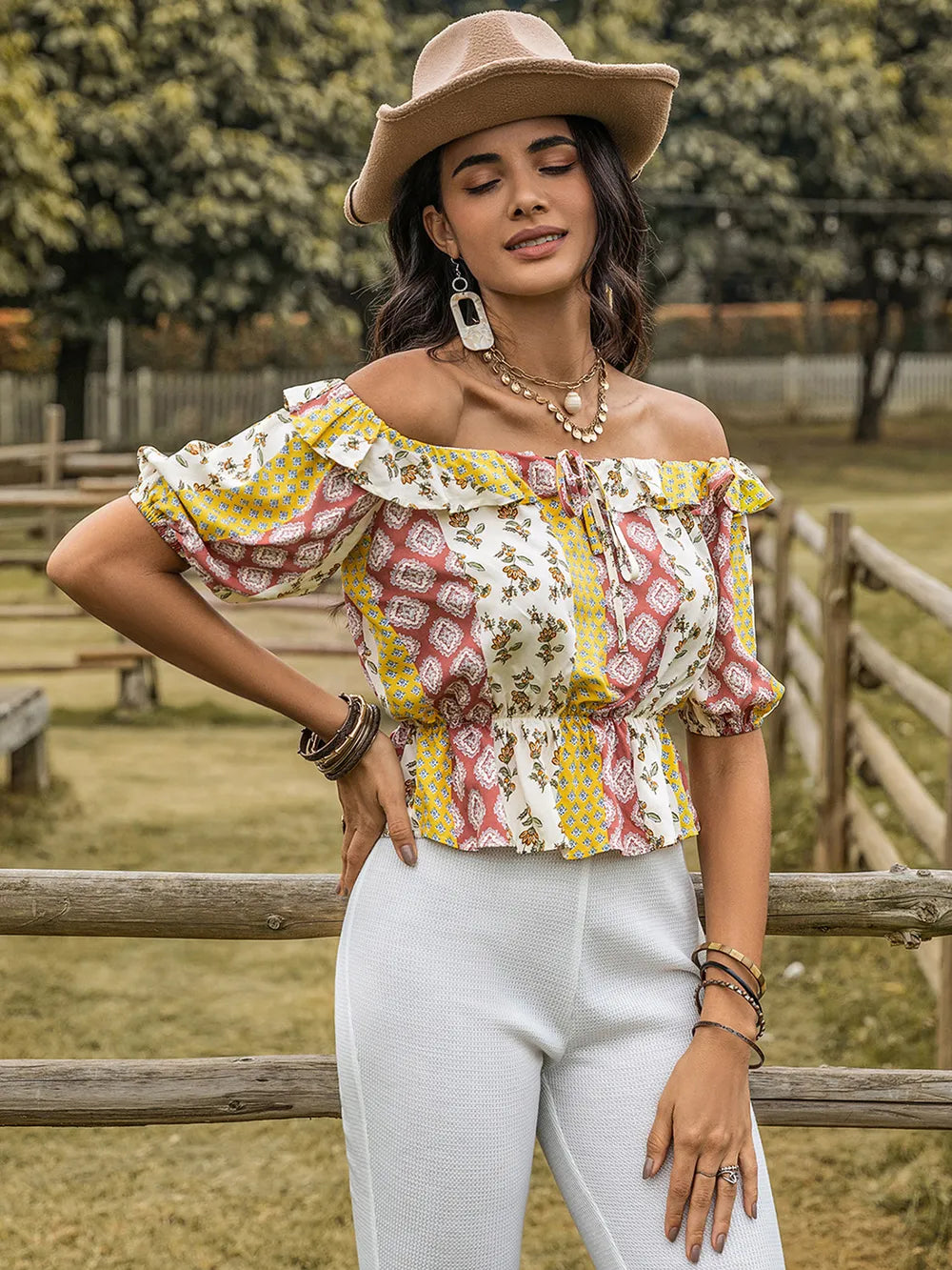 Woman wearing a colorful off-shoulder top and white pants with a hat, standing in a rustic outdoor setting.