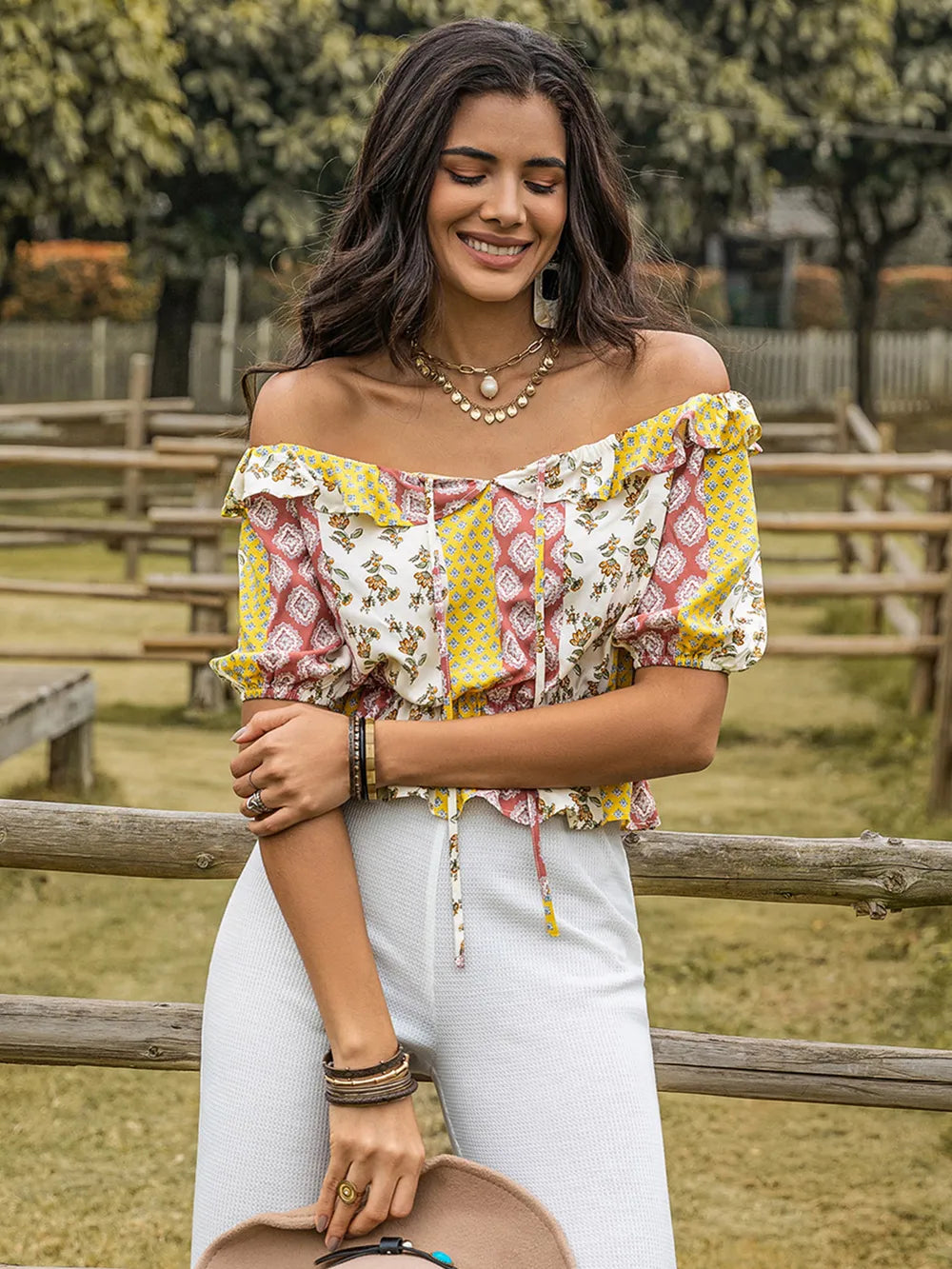 Woman wearing a colorful off-shoulder top and white pants, standing outdoors near a wooden fence.