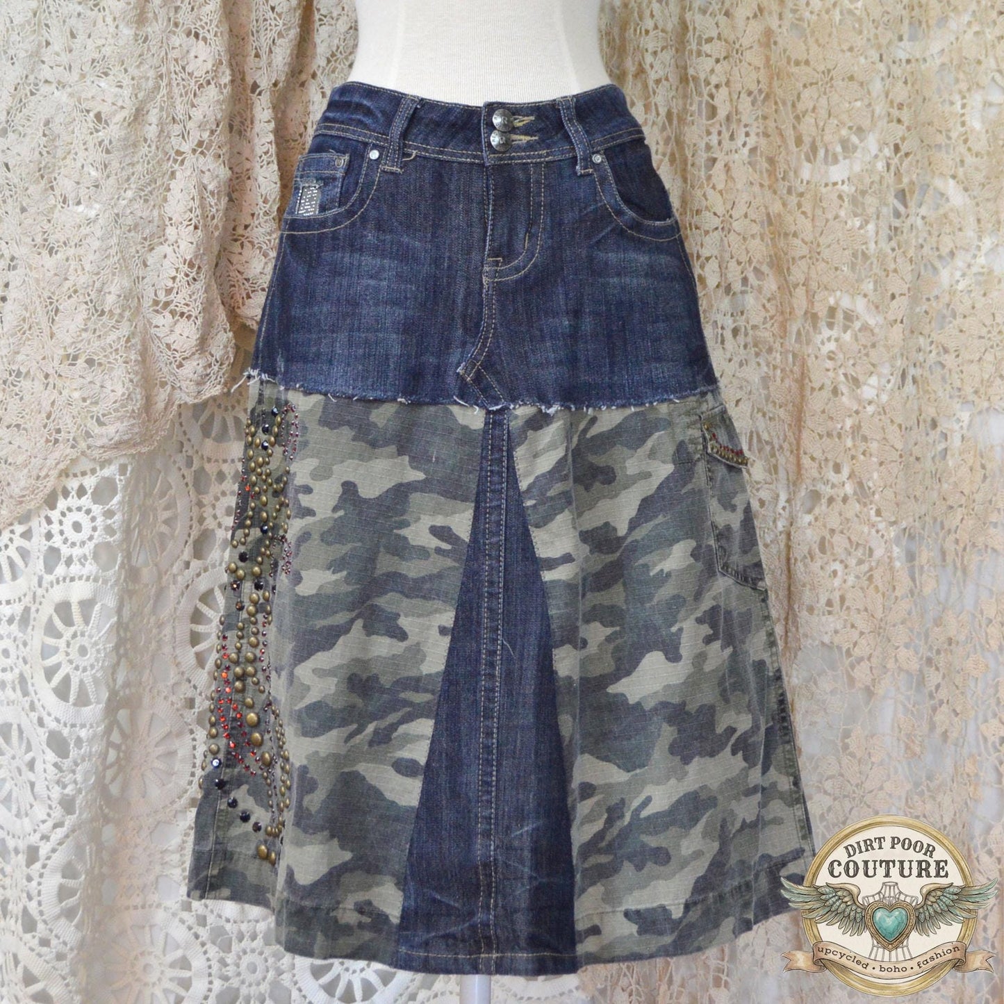 Dark wash denim skirt featuring camo fabric panels and intricate rhinestone and metal stud patterns along the front pockets and hem.