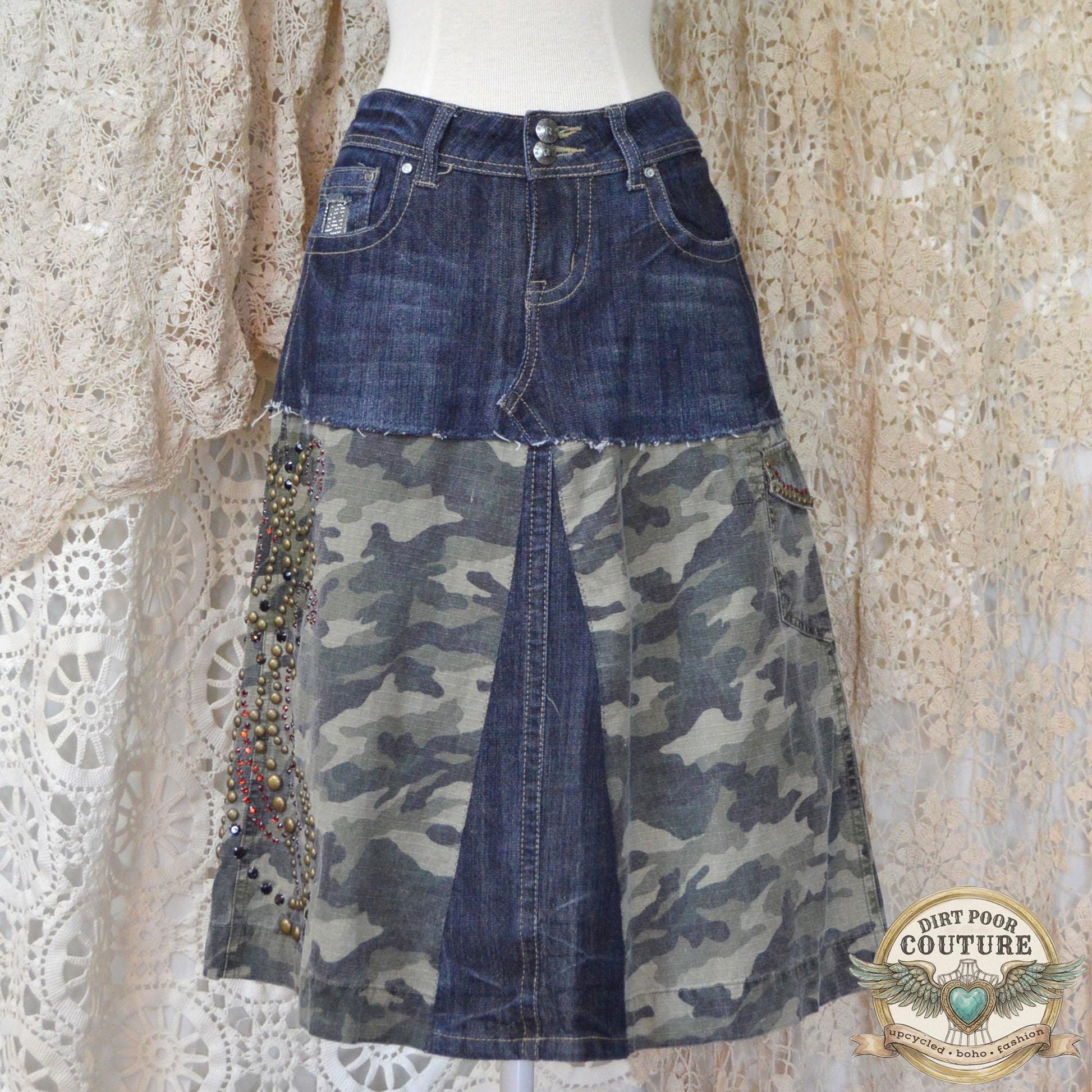 Dark wash denim skirt featuring camo fabric panels and intricate rhinestone and metal stud patterns along the front pockets and hem.