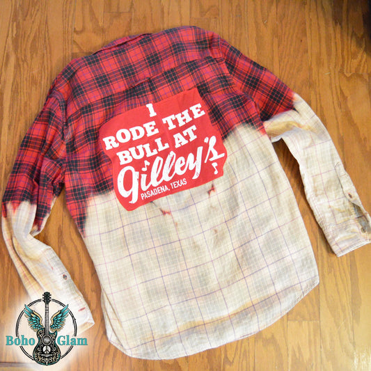 A red and black plaid flannel shirt featuring a custom dip-bleach effect that fades to a light cream color. A large red rectangular patch is sewn onto the back with white text that reads, "I RODE THE BULL AT Gilley's PASADENA, TEXAS."