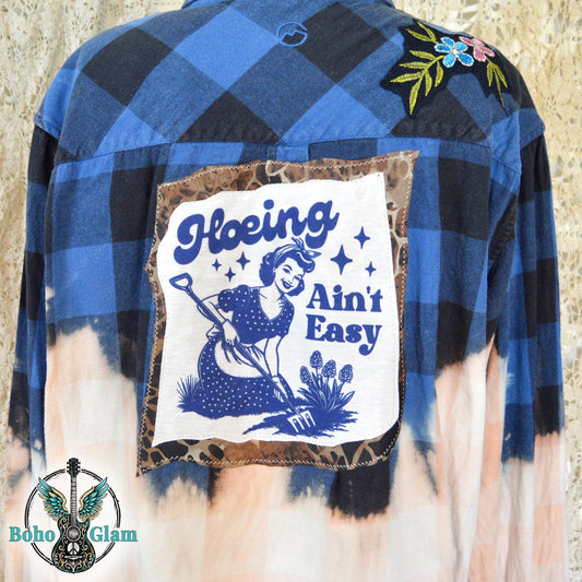 Back View Close-up: A close-up of the back of a blue buffalo plaid flannel showing a square graphic patch of a woman gardening with the text Hoeing Ain't Easy.