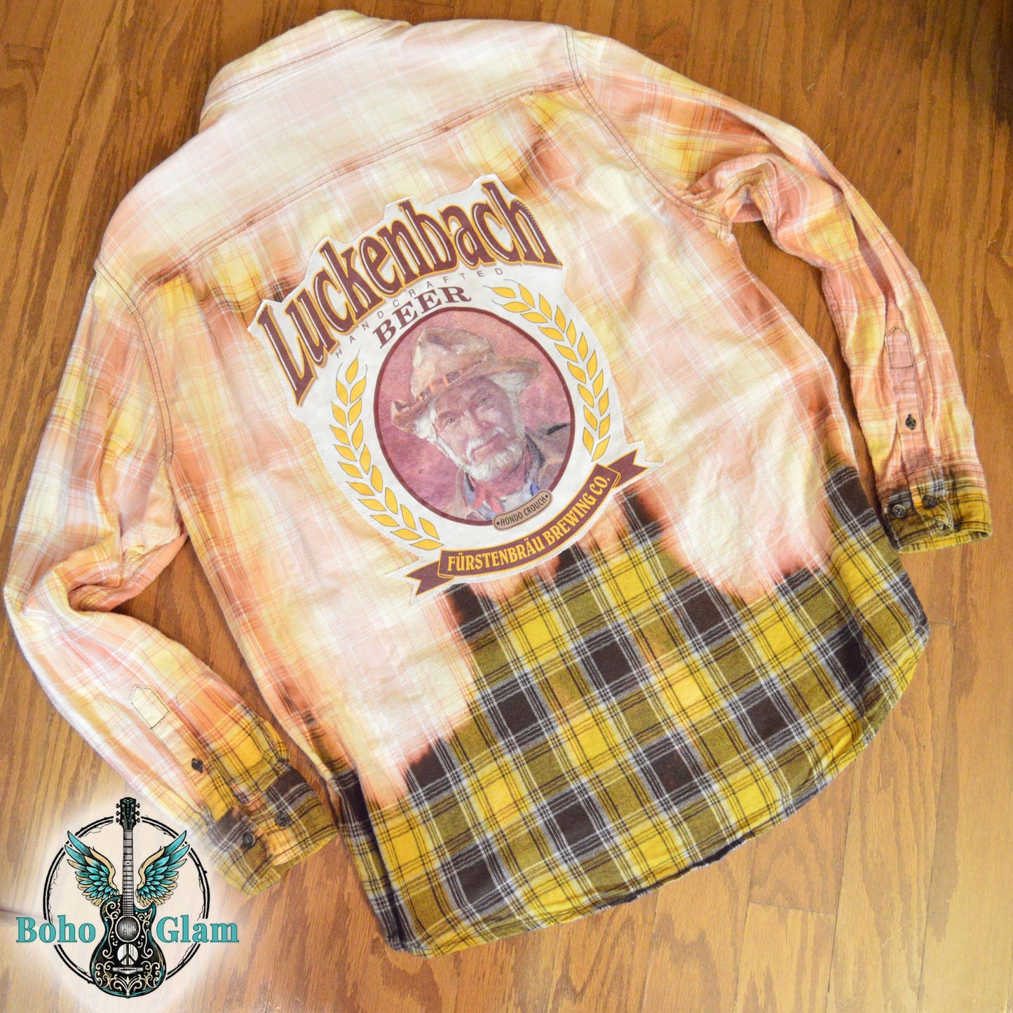 Flat Lay Back: The shirt laid flat on a wood floor to show the full scale of the Luckenbach graphic and the dip-dye bleach effect.