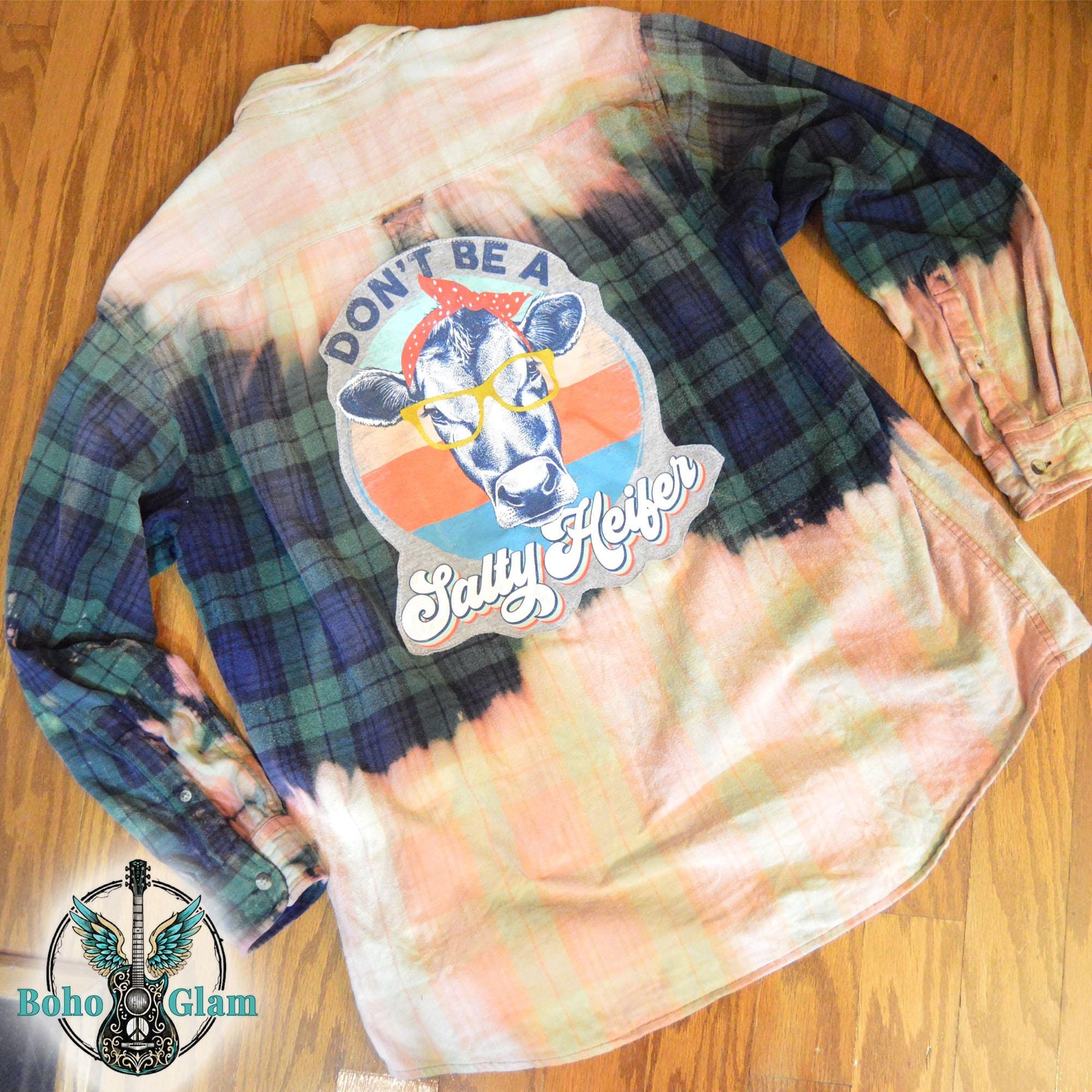 Flat Lay Back: The back of the shirt laid flat on a wood floor, highlighting the retro sunset colors of the cow graphic and the distressed bleach effect.
