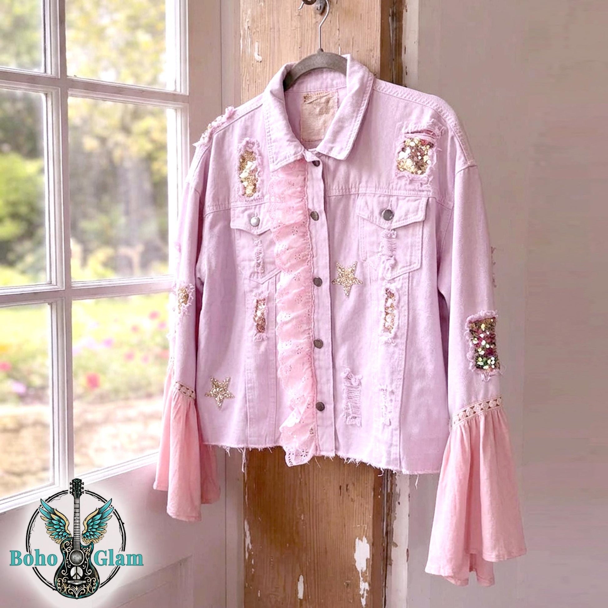 Pink upcycled denim jacket with belle sleeves, rose gold sequin patches and rhinestone stars, cropped Stevie Nicks inspired boho glam jacket, front view on hanger by window