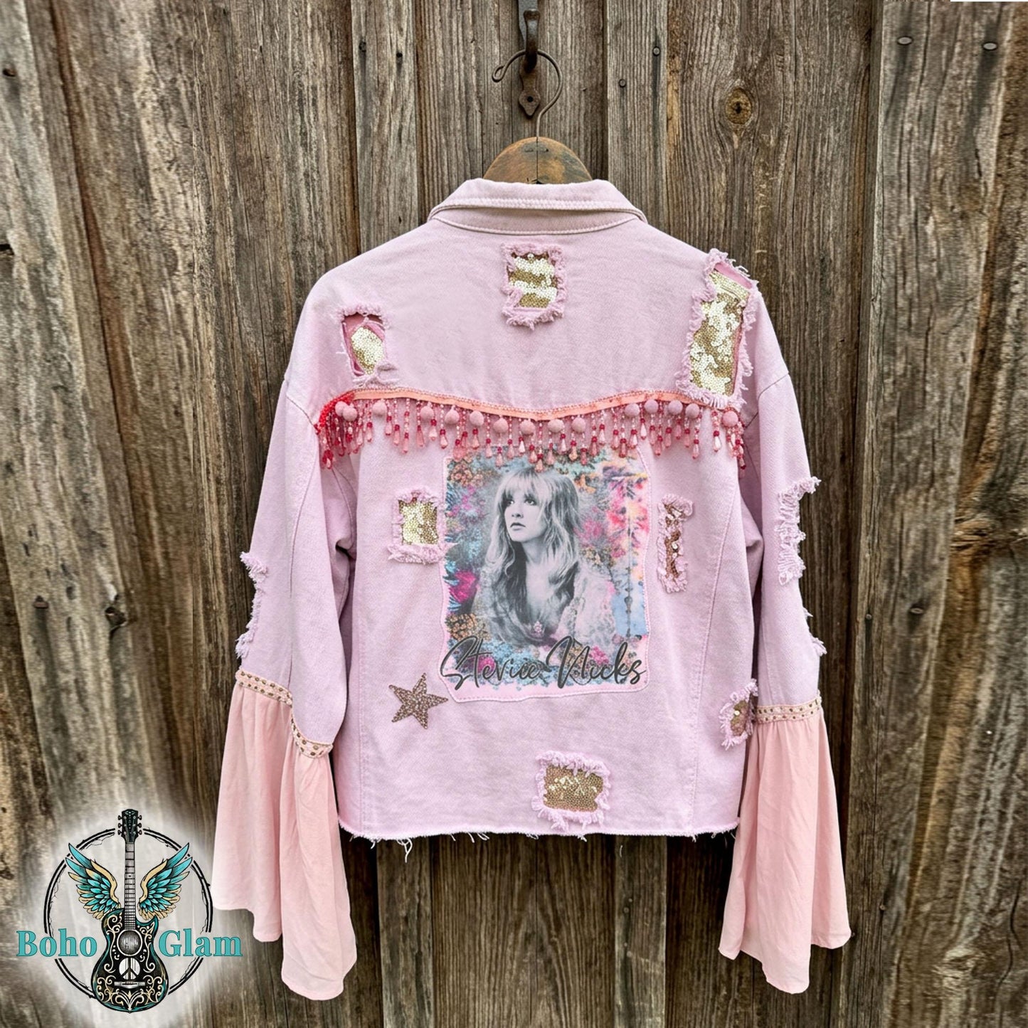 Back of pink distressed denim jacket with Stevie inspired watercolor graphic, pom trim and sequin patches, one of a kind boho glam festival jacket against rustic wood