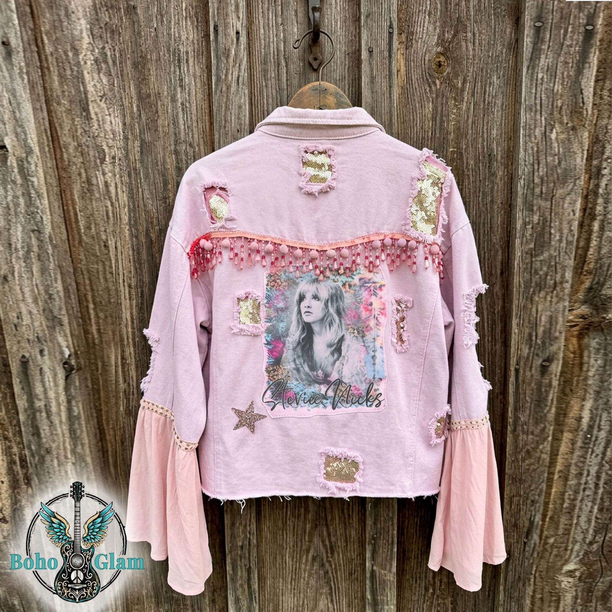 Back of pink distressed denim jacket with Stevie inspired watercolor graphic, pom trim and sequin patches, one of a kind boho glam festival jacket against rustic wood