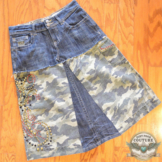 Dark wash denim skirt featuring camo fabric panels and intricate rhinestone and metal stud patterns along the front pockets and hem.