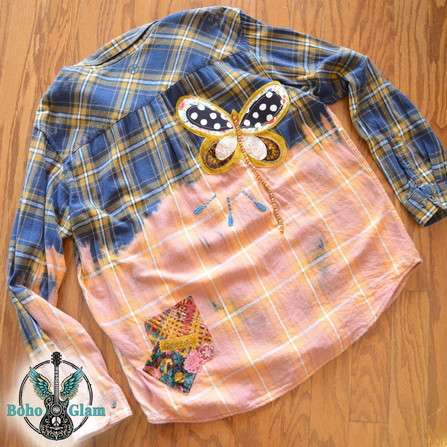 Back View: A navy and gold plaid flannel shirt with a bleached peach bottom half featuring a large butterfly applique and a 1 John 4:8 bible verse patch.