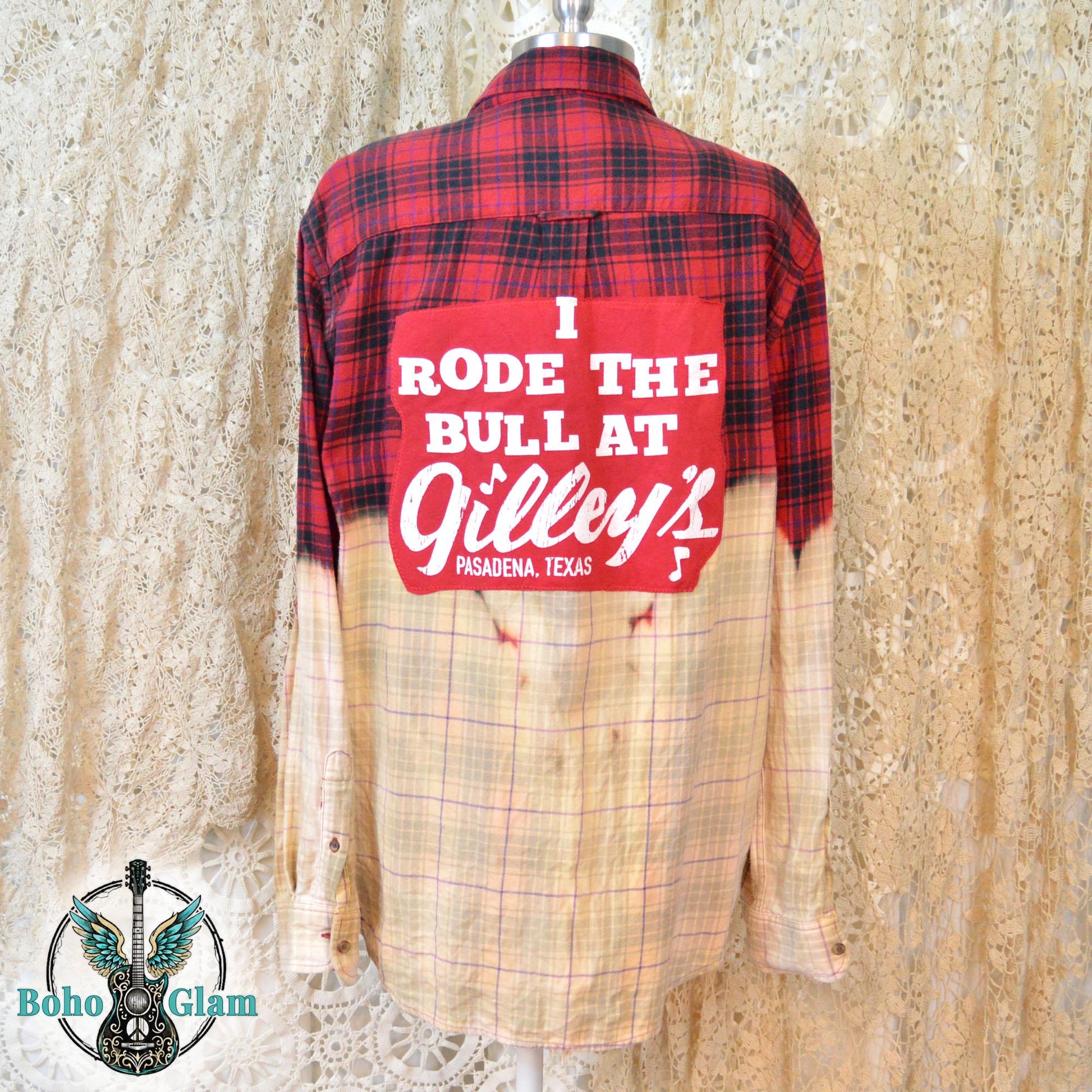 A red and black plaid flannel shirt featuring a custom dip-bleach effect that fades to a light cream color. A large red rectangular patch is sewn onto the back with white text that reads, "I RODE THE BULL AT Gilley's PASADENA, TEXAS."