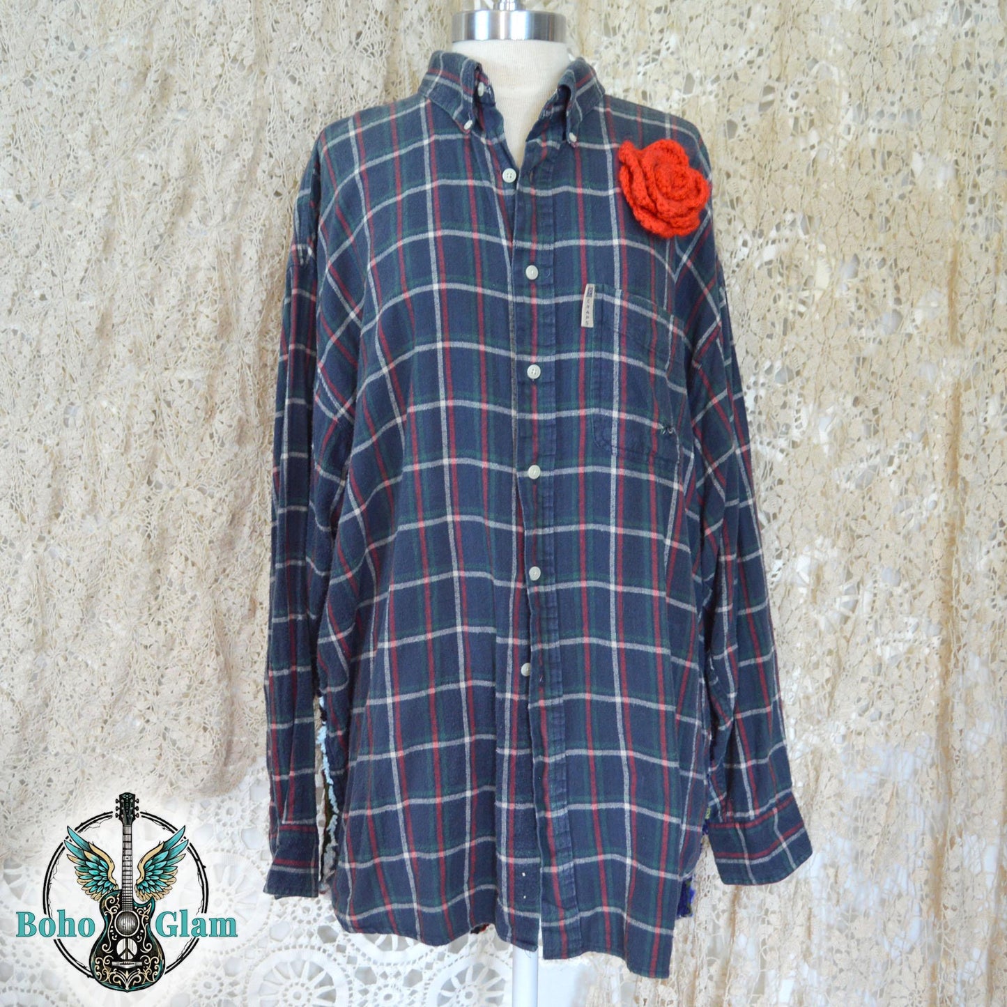 Front View on Mannequin: The front of the navy and green plaid button-down shirt featuring a prominent, handcrafted red crochet rose pinned to the upper left chest.