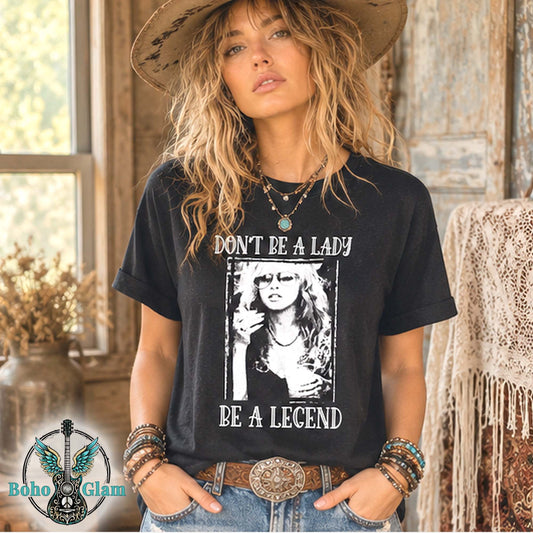 A woman wearing a black graphic t-shirt with a white print that reads 'DON'T BE A LADY BE A LEGEND' and a black-and-white photo of a woman in a hat. She is also wearing a cowboy hat, jeans, and bracelets, standing in a rustic indoor setting.